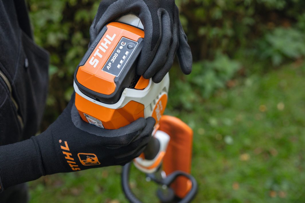 A person in black STIHL gloves inserts a STIHL AP 300 S battery into an orange and white power tool outdoors with green grass.