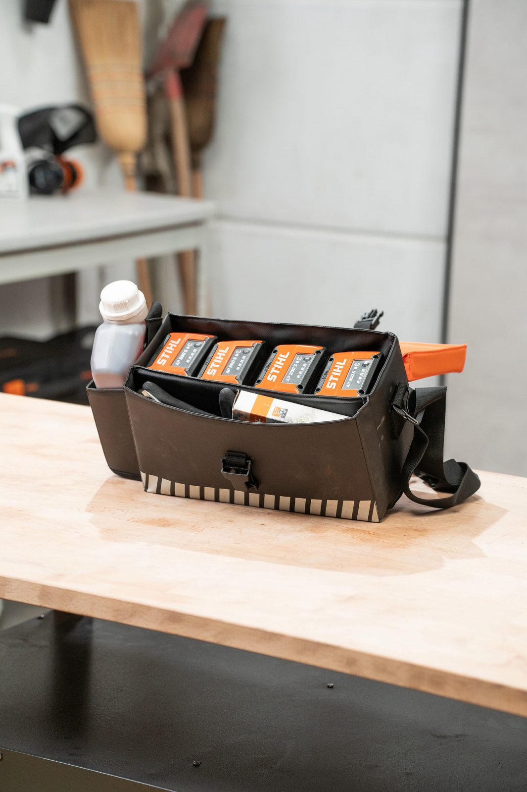 A black carry bag on a wooden workbench holds STIHL battery packs, a white bottle, an orange tool; workshop shelves in the back.