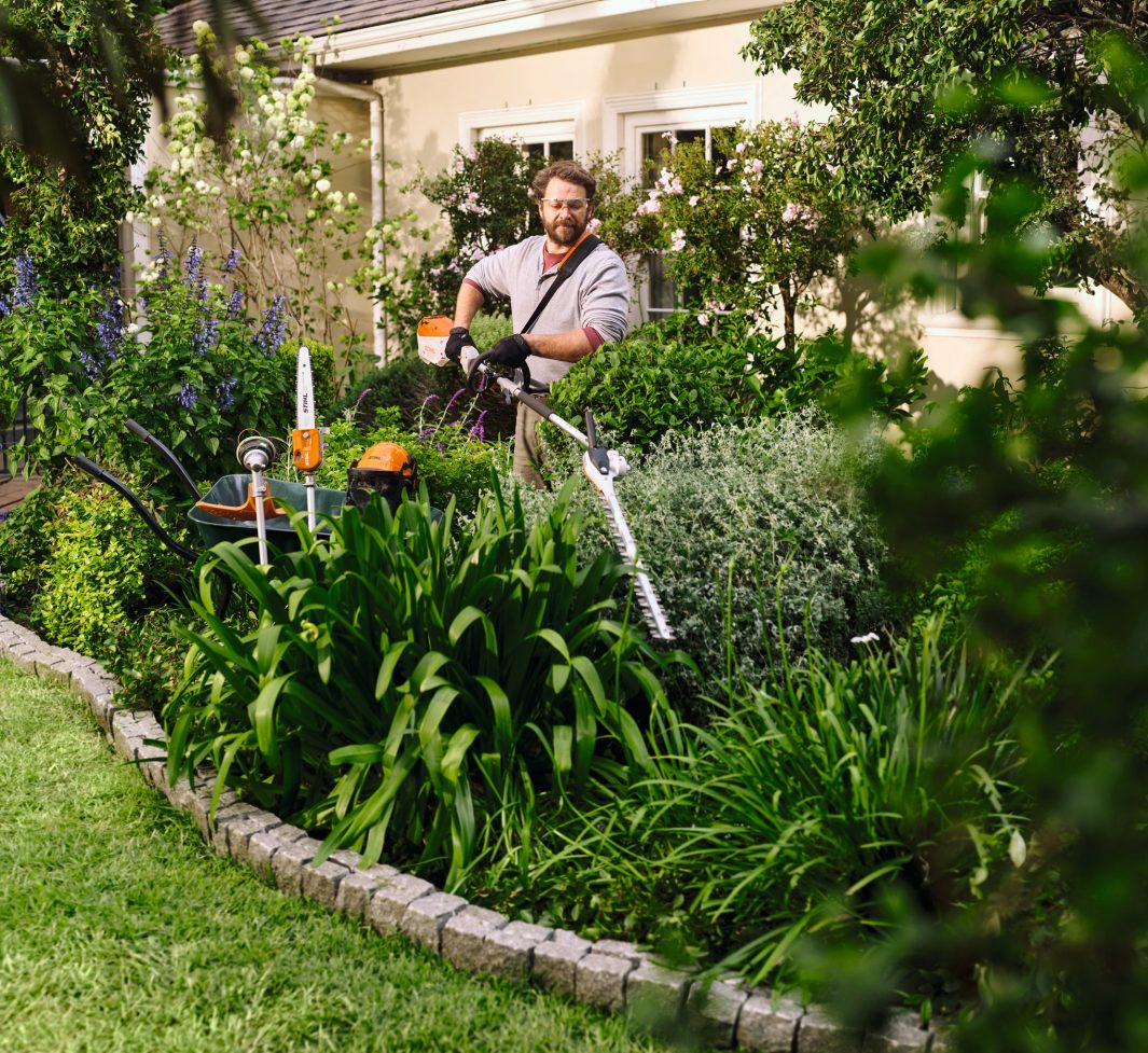 Wearing gloves and safety glasses, a man trims bushes with a Stihl KMA 80 R in a lush garden beside a house; tools in foliage.