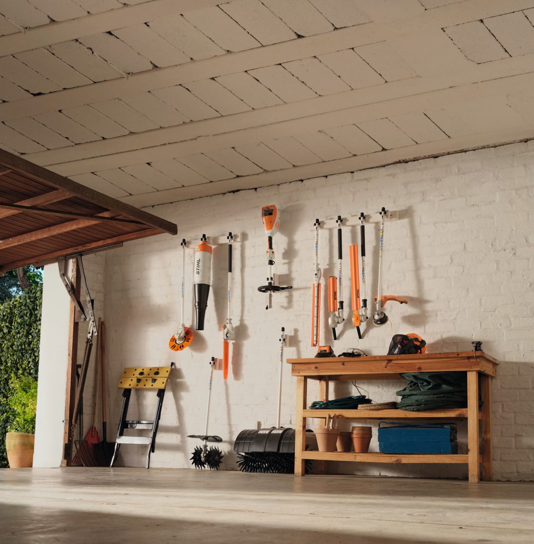 A tidy garage with garden tools on a white brick wall, workbench with pots and cloth, and open door revealing greenery outside.
